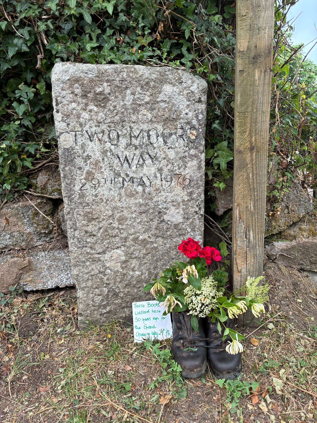 Two Moors Way marker stone at Morchard Bishop with walking boots and flowers